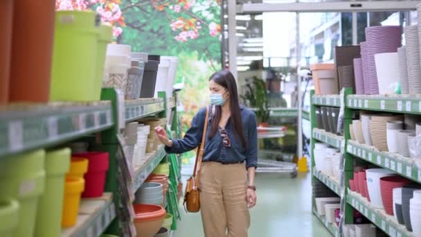 Jonge vrouwelijke klant in het gezicht masker op zoek naar planken met veel verschillende bloempotten - Video