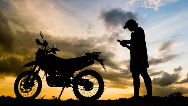 The silhouette of an adventurous traveler man looking at mobile phone with motocross motorcycle in the evening - Foto, Imagem