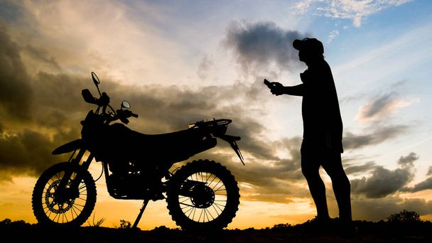 Sylwetka śmiałego podróżnika patrzącego wieczorem na telefon komórkowy z motocyklem motocross - Zdjęcie, obraz