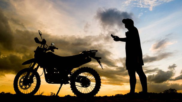 The silhouette of an adventurous traveler man looking at mobile phone with motocross motorcycle in the evening - Photo, Image