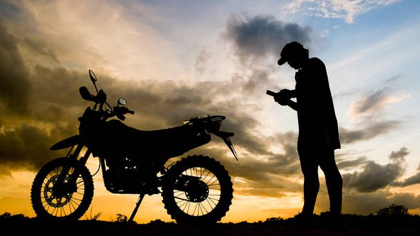 Sylwetka śmiałego podróżnika patrzącego wieczorem na telefon komórkowy z motocyklem motocross - Zdjęcie, obraz