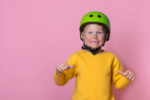 Charming blond kid boy in bright green helmet and yellow clothes with thumbs up pointing on him. School kid isolated on pink. Funny face. Portrait of stylish 7 years old kid. Space for text on shirt - Fotoğraf, Görsel