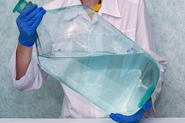 pracownik medyczny w białym płaszczu i niebieskich rękawiczkach ochronnych posiada ogromną butelkę płynnych leków koncepcyjnych szczepień. Wysokiej jakości zdjęcie - Zdjęcie, obraz