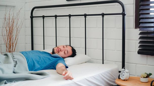 A fiatal ázsiai férfi reggel felébred, ázsiai férfi mobilon beszél a barátaival, miután felébredt az ágyában otthon, a hálószobában. Jóképű férfiak nap, álmos pihenés a modern ház koncepció. - Fotó, kép