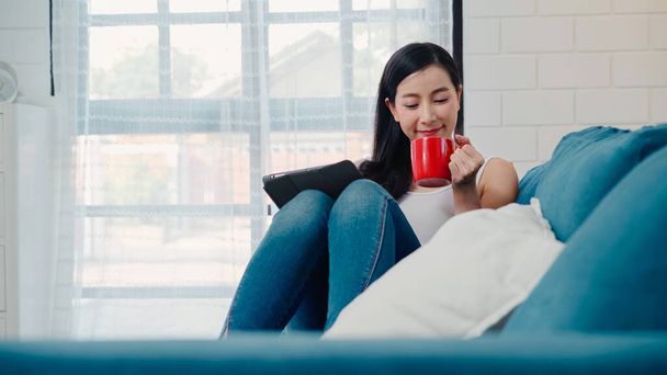 Beautiful attractive smiling Asian woman using tablet holding a warm cup of coffee or tea while lying on the sofa when relax in living room at home. Lifestyle women at home concept. - Photo, Image