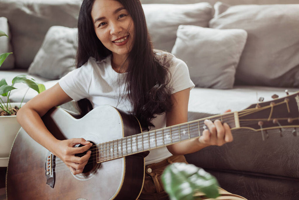 Mujer tocando la guitarra en casa. Mujer hermosa sonriendo y tocando la guitarra con sus plantas en la sala de estar. - Foto, imagen
