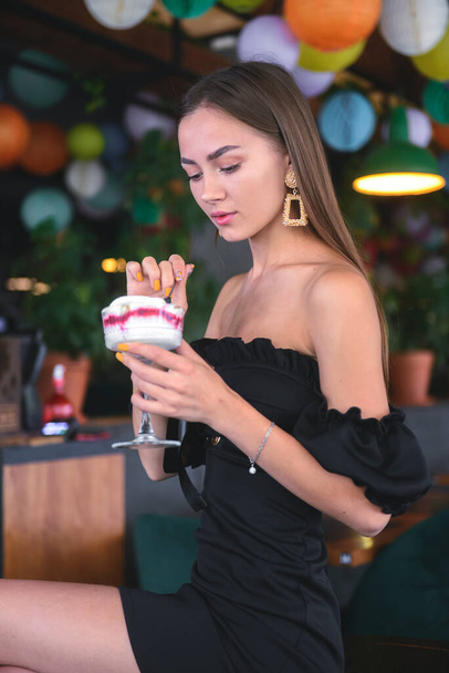 Beautiful sexy brunette woman in restaurant or cafe with ice cream tiramisu dessert. Fashion photo of young woman in black dress with hairstyle and makeup, eating dessert. Eat out concept. - Foto, afbeelding