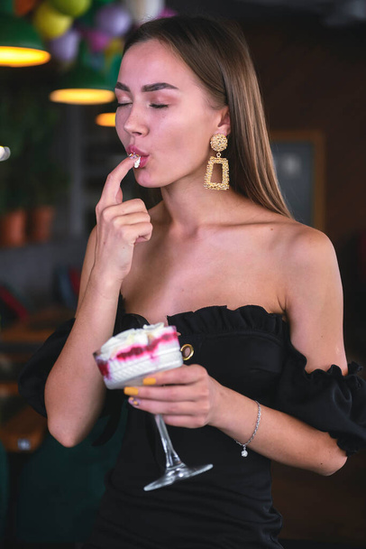 Beautiful sexy brunette woman in restaurant or cafe with ice cream tiramisu dessert. Fashion photo of young woman in black dress with hairstyle and makeup, eating dessert. Eat out concept. - Zdjęcie, obraz