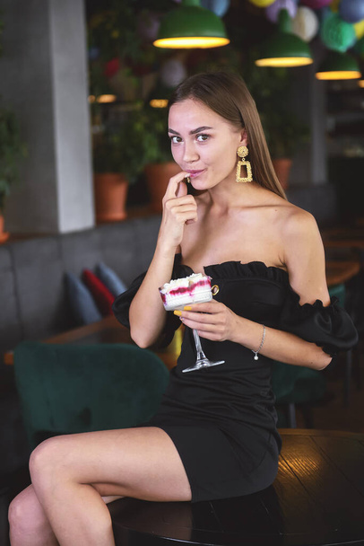 Beautiful sexy brunette woman in restaurant or cafe with ice cream tiramisu dessert. Fashion photo of young woman in black dress with hairstyle and makeup, eating dessert. Eat out concept. - Фото, зображення