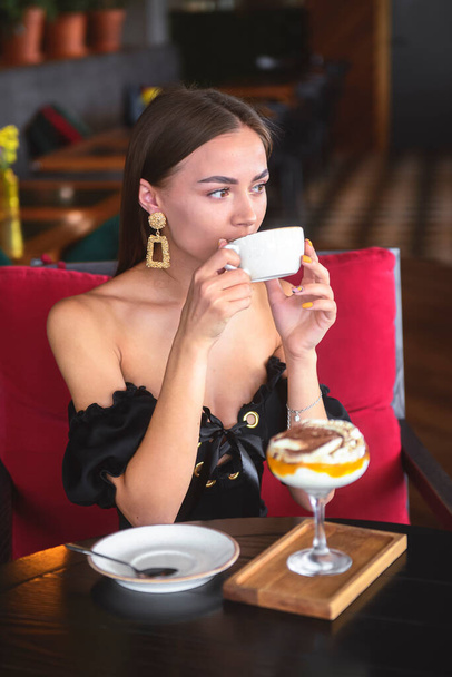Beautiful sexy brunette woman in restaurant or cafe with ice cream tiramisu dessert and a cup of . Fashion photo of young woman in black dress with hairstyle and makeup eat out concept. - Photo, Image