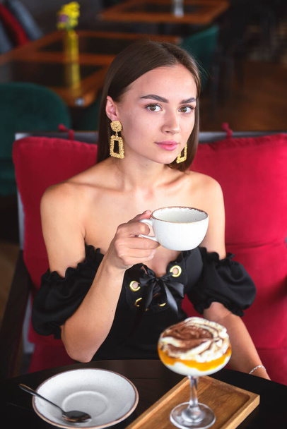 Beautiful sexy brunette woman in restaurant or cafe with ice cream tiramisu dessert and a cup of . Fashion photo of young woman in black dress with hairstyle and makeup eat out concept. - Foto, Bild