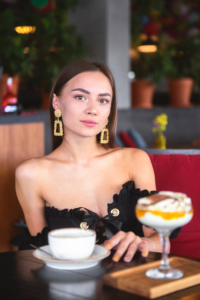Beautiful sexy brunette woman in restaurant or cafe with ice cream tiramisu dessert and a cup of . Fashion photo of young woman in black dress with hairstyle and makeup eat out concept. - Фото, изображение