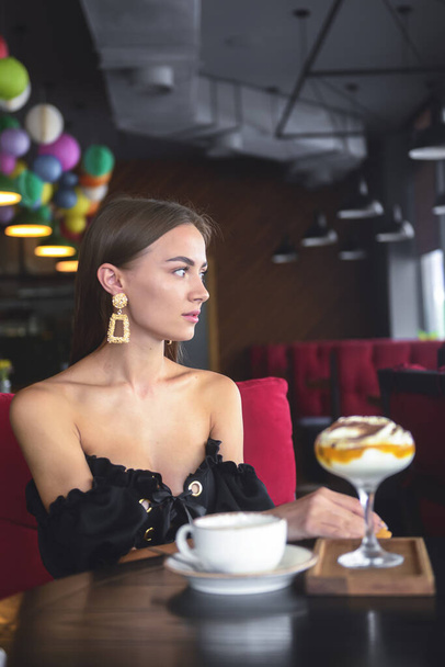 Beautiful sexy brunette woman in restaurant or cafe with ice cream tiramisu dessert and a cup of . Fashion photo of young woman in black dress with hairstyle and makeup eat out concept. - Photo, image