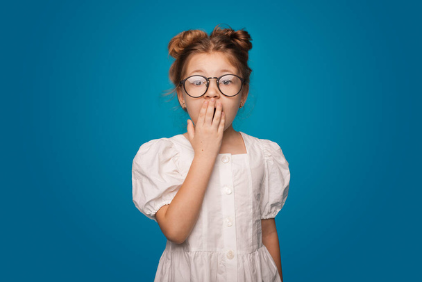 Überraschtes kleines Mädchen bedeckt den Mund und posiert in einem Kleid auf einer blauen Studiowand - Foto, Bild