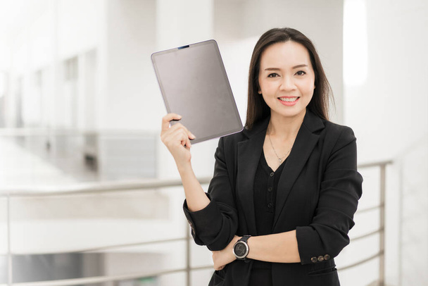 Portrait stock photo of a confident cheerful Asian businesswoman professional in black business suit using a digital tablet standing in the office building representing business oriented concept - Photo, image