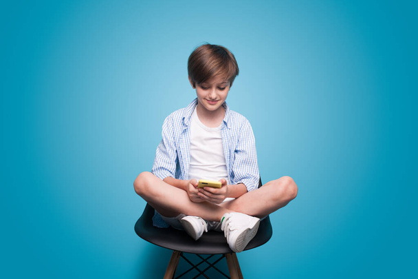 Beyaz bir öğrenci koltukta oturuyor ve mavi bir duvardaki telefonu kullanıyor. - Fotoğraf, Görsel