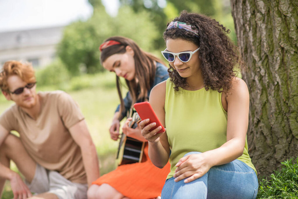 Girl looking into smartphone girlfriend with guitar and boyfriend - Zdjęcie, obraz