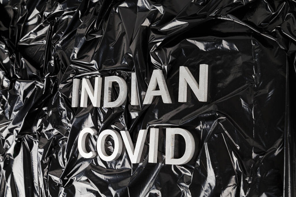 palabras indio covid laid with silver metal letters on crumpled black plastic bag background in slanted perspective - Foto, Imagen