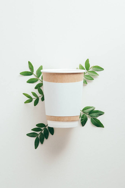 Take-out cardboard coffee cup mockup with green leaves on white background. Coffee cup to go design, branding - Фото, изображение