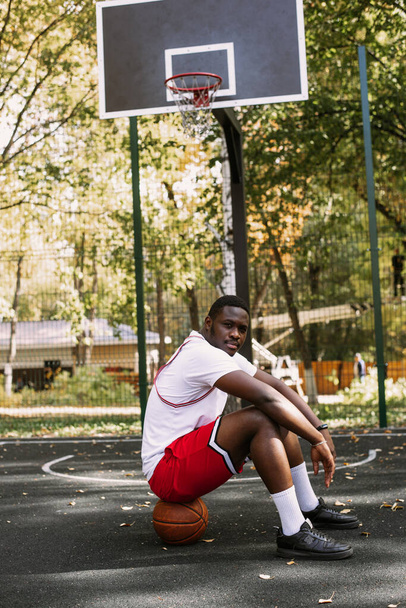 Elinde basketbol topu olan ve basketbol sahasında oturan Afro-Amerikan bir gencin portresi. Egzersiz yaparken biraz ara ver. Modaya uygun spor portresi - Fotoğraf, Görsel