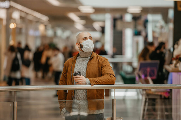 コロナウイルスの拡散を避けるため顔マスクの男がショッピングセンターで待っている間にコーヒーカップを持っている。外科用マスクのはげ男は社会的距離を保っている. - 写真・画像