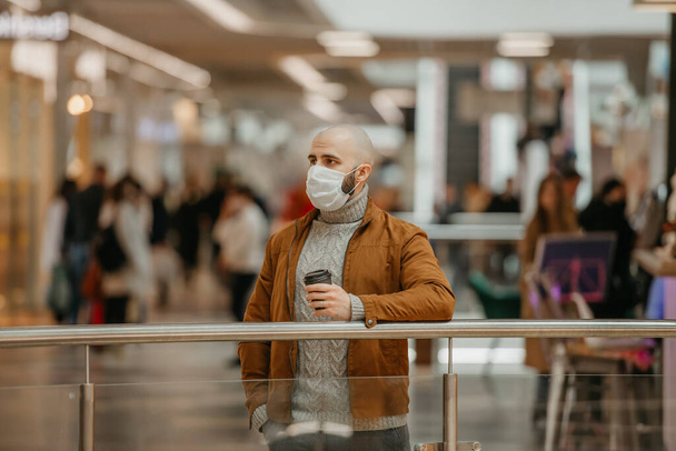 Un hombre con una máscara facial médica para evitar la propagación del coronavirus está sosteniendo una taza de café mientras espera en el centro comercial. Un tipo calvo con una máscara quirúrgica mantiene una distancia social.. - Foto, imagen