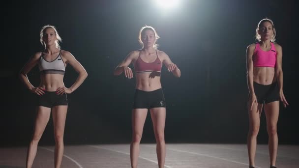 Three women athletes prepare for a track race in a dark stadium with streetlights on. Time-lapse footage of warm-up and concentration of a group of women before the race on the track - Filmagem, Vídeo