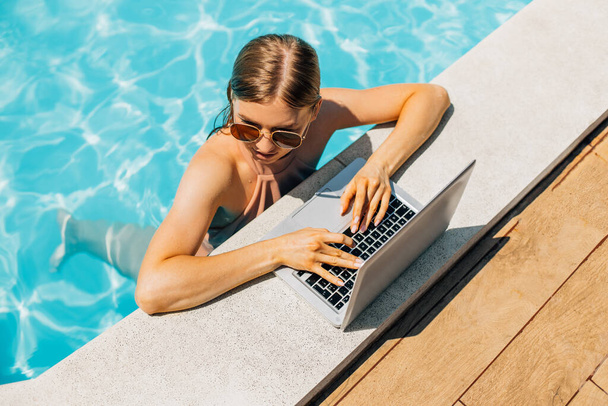 Vonzó fiatal nő napszemüvegben, laptopot használ és távolról dolgozik a medence felett, vonzó nő mosolyog, laptopot használ a medencében, nyári vakáció koncepció, távoli munka - Fotó, kép