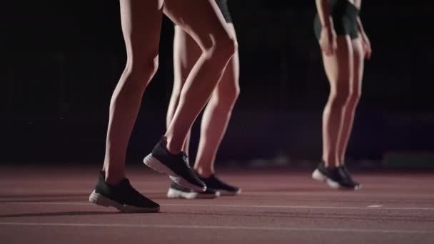 Drie vrouwelijke atleten bereiden zich voor op een baanrace in een donker stadion met straatlantaarns aan. Time-lapse beelden van warming-up en concentratie van een groep vrouwen voor de race op het circuit - Video