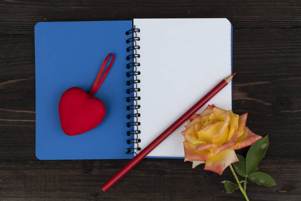 On a dark wooden background lies an open notebook, on one page of which is a red heart, and on the other - a rose and a red pencil. - Photo, image
