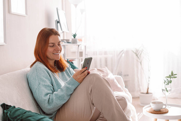 Happy beautiful girl using mobile phone, sitting in the living room. - Photo, Image