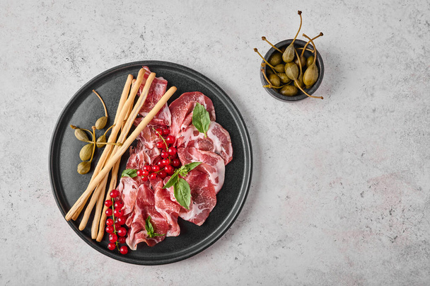 Slices of prosciutto coppa di parma ham with basil, grissini sticks, capers and redcurrant on dark plate - Photo, image