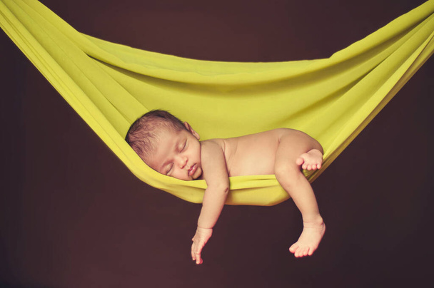 15 ημέρες νεογέννητο μωρό κοιμάται σε μια φωτεινή κίτρινη αιώρα, κρέμονται ύφασμα με το χέρι και το πόδι της κρέμεται έξω. νεογέννητο πορτρέτο - Φωτογραφία, εικόνα