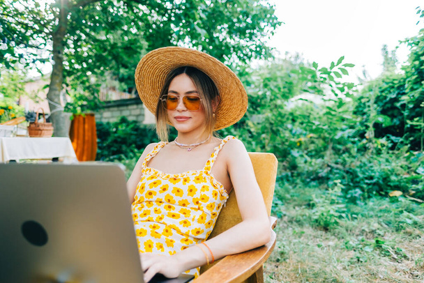 Young caucasian woman using laptop computer outdoor, sitting on a bench in the backyard in summer day. - Foto, Imagen