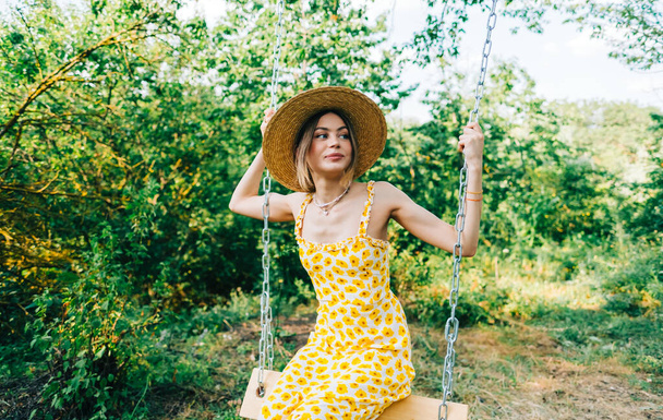 Portrait of attractive young woman in straw hat outdoors in summer sunny day on a swing. - Foto, imagen