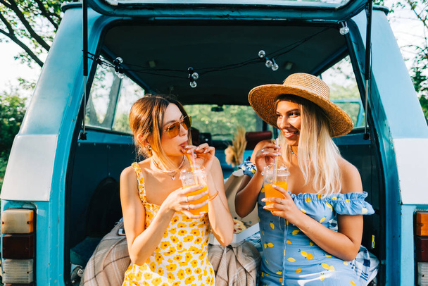 Duas mulheres atraentes e alegres bebendo limonada perto de van, desfrutando de vibrações de verão em viagem de carro - Foto, Imagem