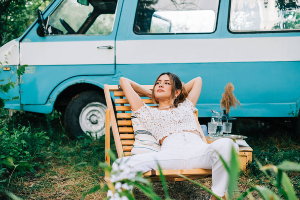 Young woman resting in nature, sitting on wooden chair near van outdoors. Enjoying summer, travel concept - Фото, зображення