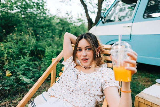 Portrait of young woman sitting on wooden chair near van and drinking lemonade outdoors in nature. Enjoying summer, travel concept - Fotoğraf, Görsel