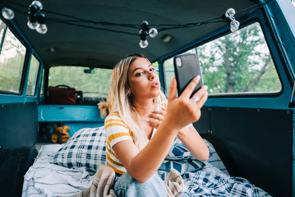 Young woman sitting in a van and using mobile phone, looking on screen, outdoors in nature. Enjoying summer, travel concept - Фото, изображение