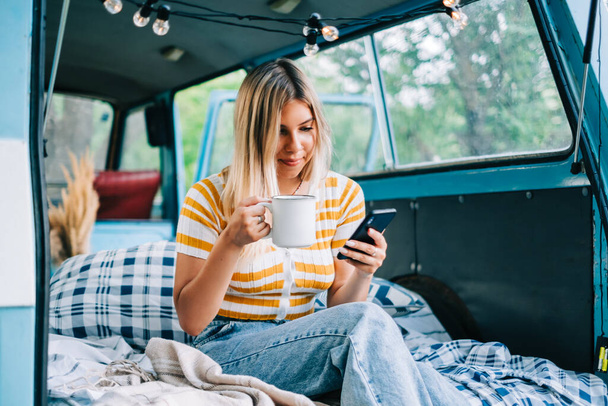 Portrait of young woman sitting in a van and using mobile phone, outdoors in nature. Enjoying summer, travel concept - Photo, Image