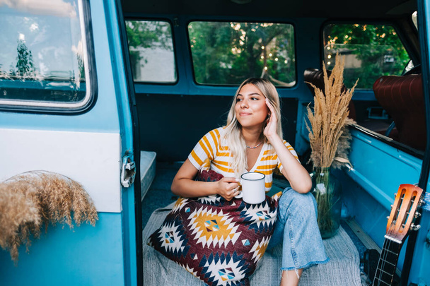 Portrait of happy young woman sitting in a van and drinking tea outdoors in nature, during sunset. Enjoying summer, travel concept - Photo, image