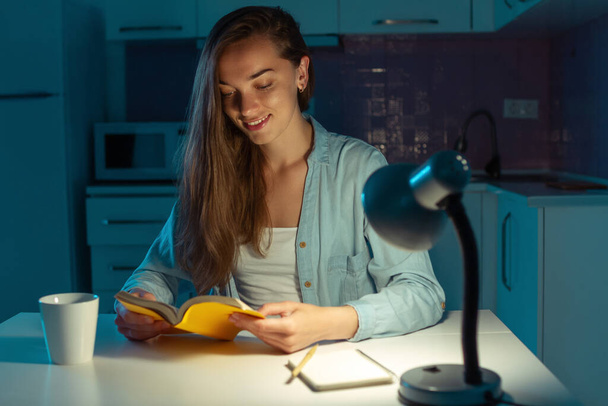 Portrait of young, attractive, smiling woman reading a book in the evening at home at the kitchen. Night reading - Foto, immagini