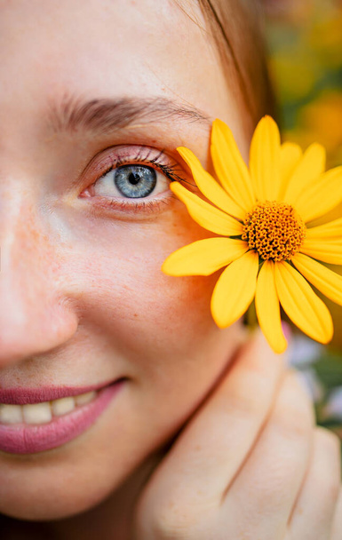 Portrait Of Young Woman With Blue Eyes Holding A Yellow Flower Over Her Eye. - 写真・画像