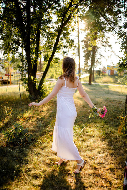 Egy nő a parkban, aki aranyos ruhát visel, virágot tart egy meleg nyári napon, naplementekor.. - Fotó, kép