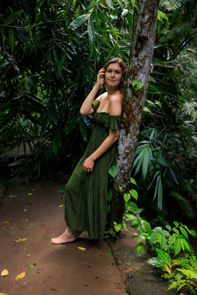 Caucasian woman leaning against a tree in tropical rain forest. Beautiful woman wearing long green dress. Walking trail in jungle. Nature concept. Travel to Asia. Bali island, Indonesia - Photo, Image