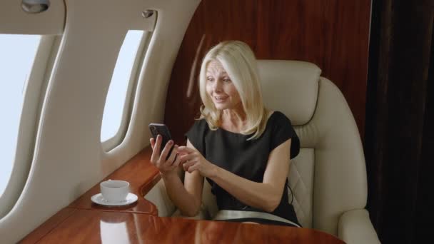 Excited mature female looks at smartphone and rejoices in victory while flying airplane. Happy gray-haired rich senior woman celebrating success win on mobile phone while traveling in private jet - Footage, Video
