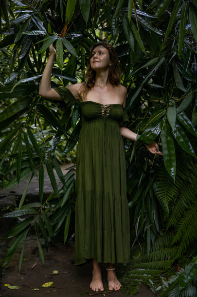 Beautiful woman walking in tropical rain forest, wearing long green dress. Woman in jungle touching bamboo leaves. Nature and environment concept. Travel to Asia. Bali island, Indonesia - Photo, Image