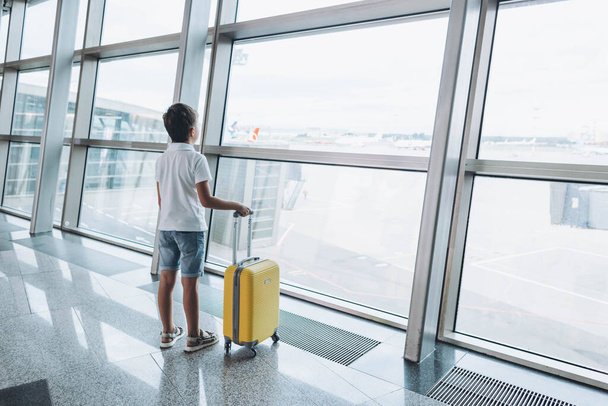 空の空港ターミナルで黄色のスーツケースを着た男の子が窓の外を見て出発を待っています。Tシャツとショートパンツの子供たちは、飛行機のフライトを待っているラウンジに立っています。家族旅行のコンセプト - 写真・画像
