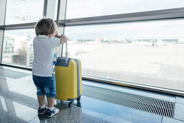 Boş havaalanı terminalinde sarı valizli küçük bir çocuk. Tişörtlü ve şortlu çocuk salonda uçak uçuşunu bekliyor. Aile gezisi ve tatil konsepti. - Fotoğraf, Görsel