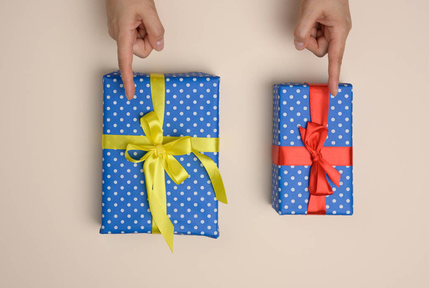 дві подарункові коробки, загорнуті в шовкову стрічку на бежевому фоні, жіночі пальці вказують на вибір
 - Фото, зображення